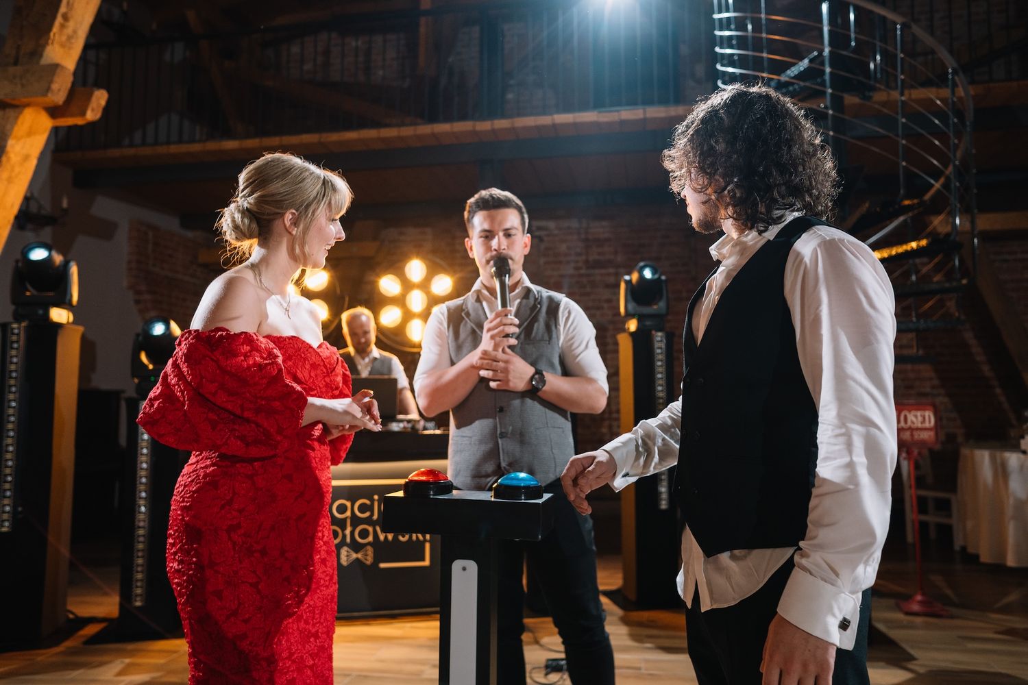 Woman in red dress and man with curly hair interact with host at wedding reception game.