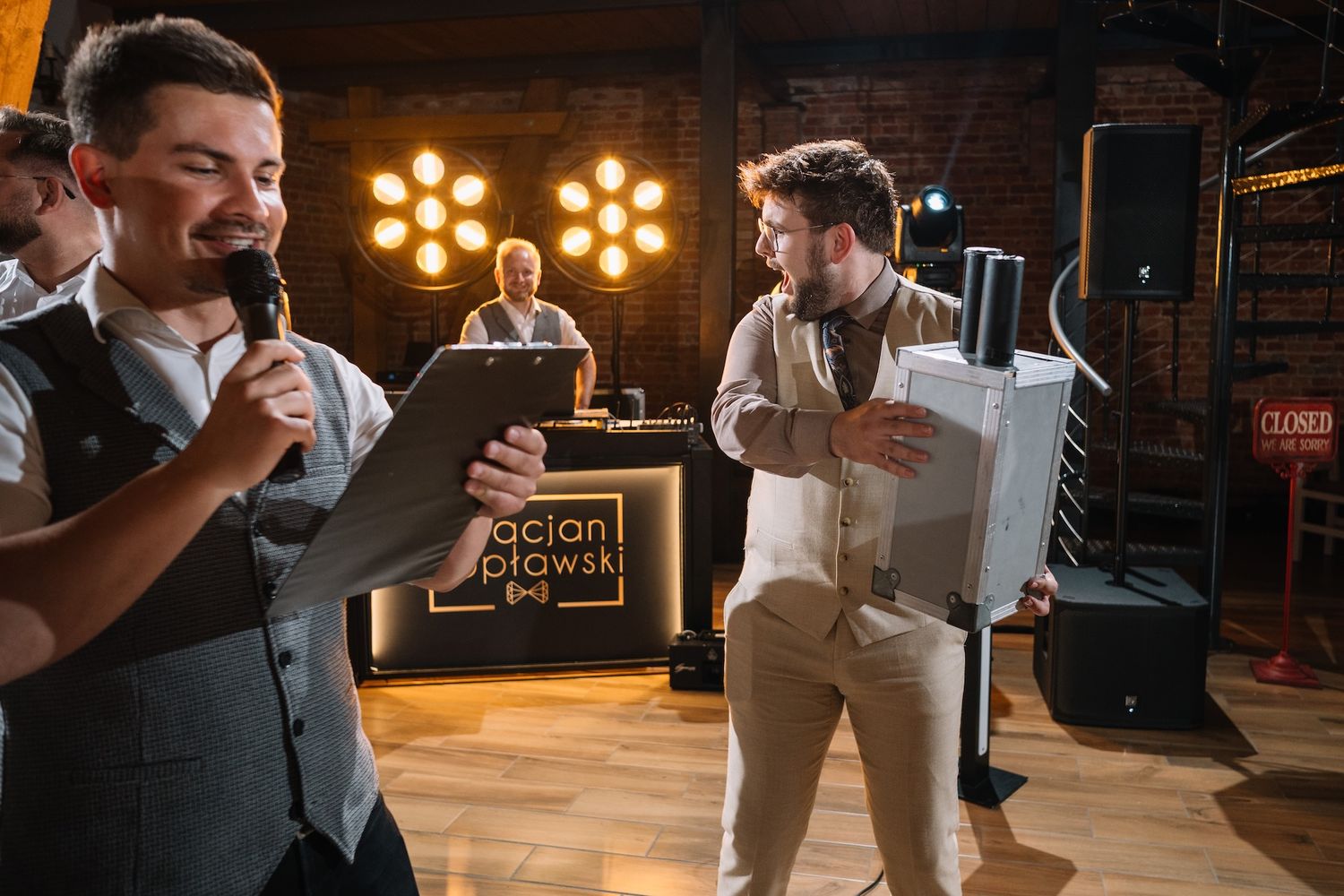 Wedding MCs with microphone and clipboard host entertainment segment at rustic barn reception.
