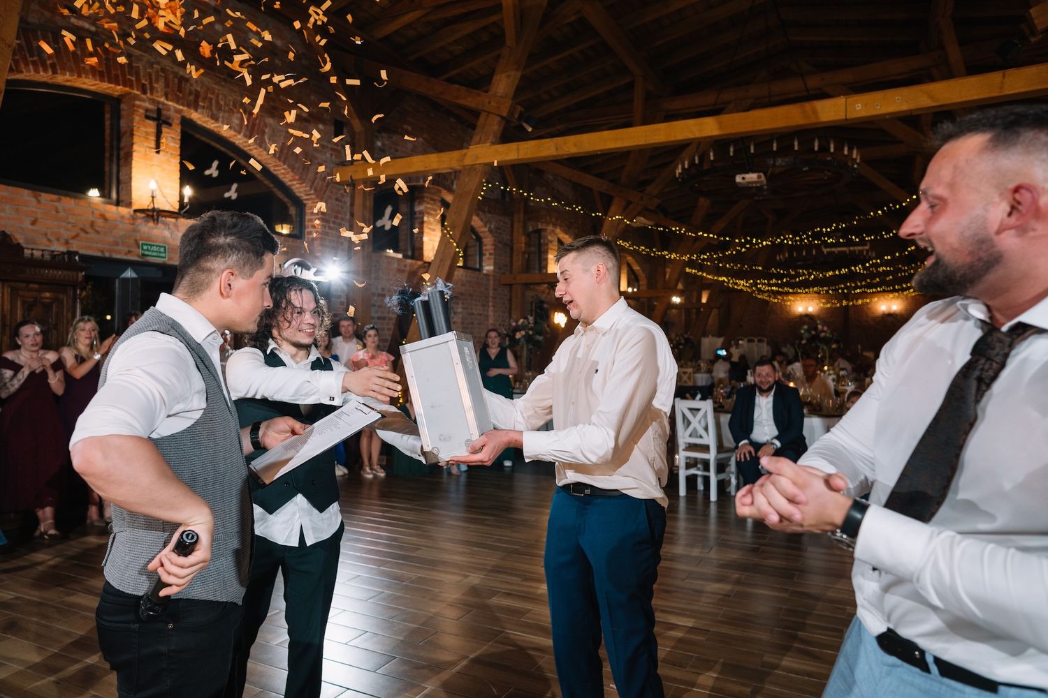 Groom receiving a gift from guests during wedding reception in a rustic barn venue with golden confetti.