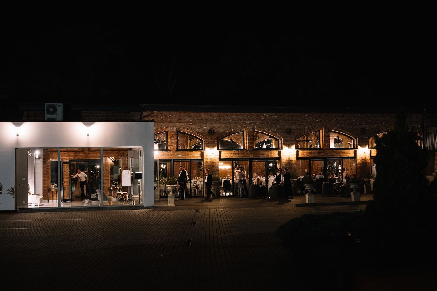 Rustic wedding venue exterior illuminated at night with warm lights and guests gathered outside.
