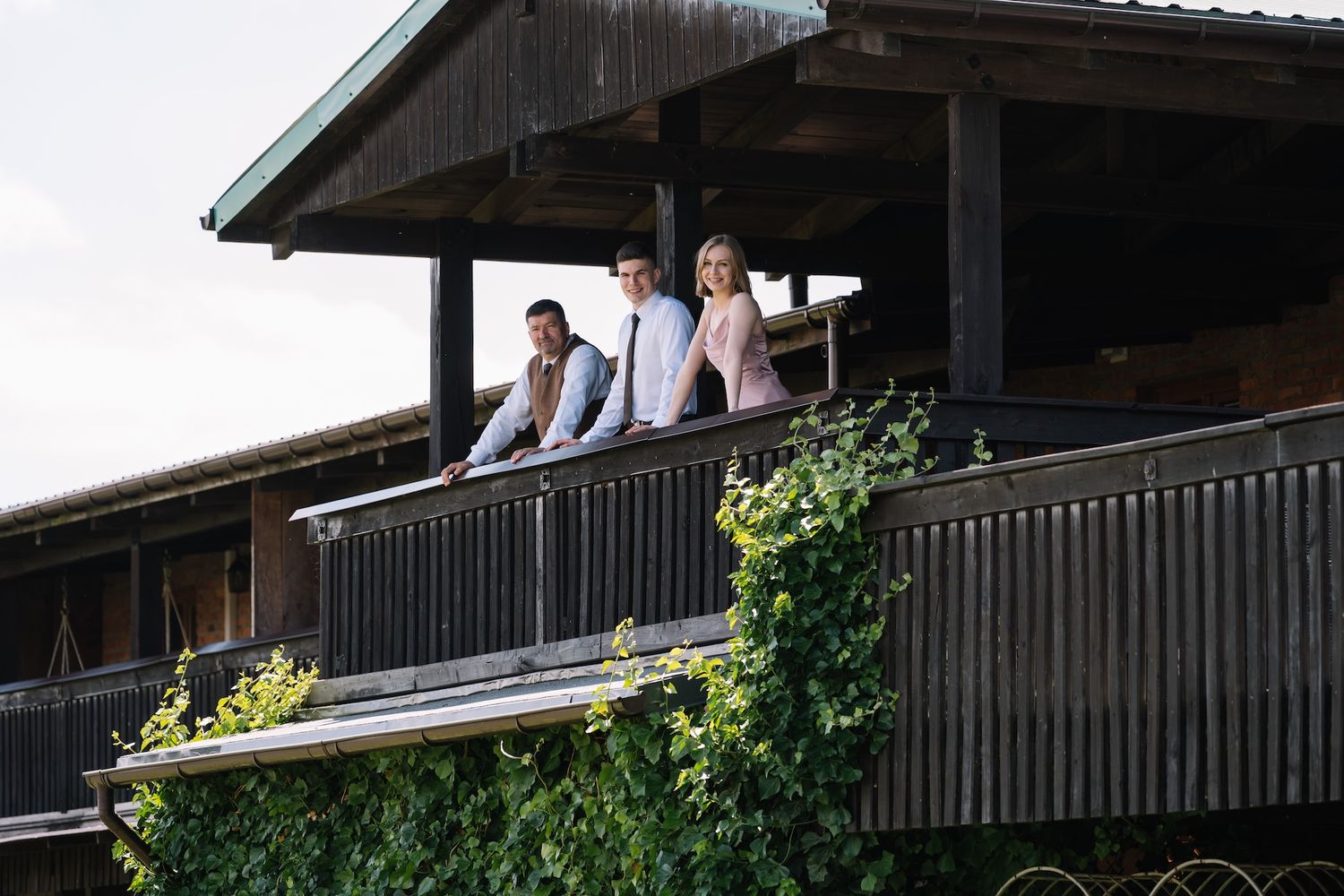 Three elegantly dressed wedding guests pose on a rustic wooden balcony with green ivy.