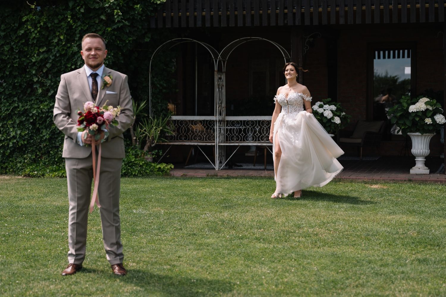 Groom in gray suit holds bouquet as bride in white gown approaches across green lawn.