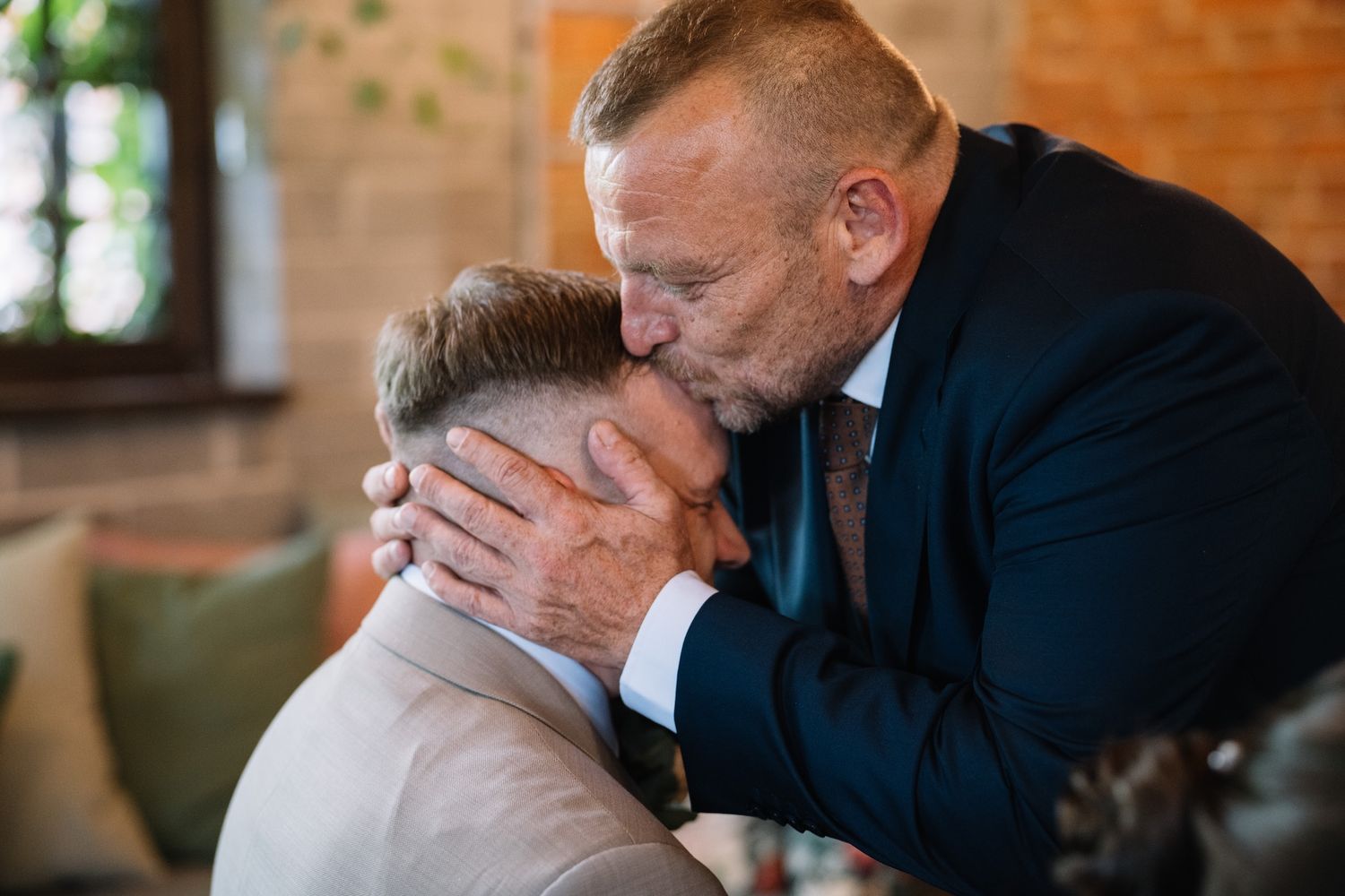 Older man tenderly kisses groom on forehead in emotional pre-wedding moment indoors.