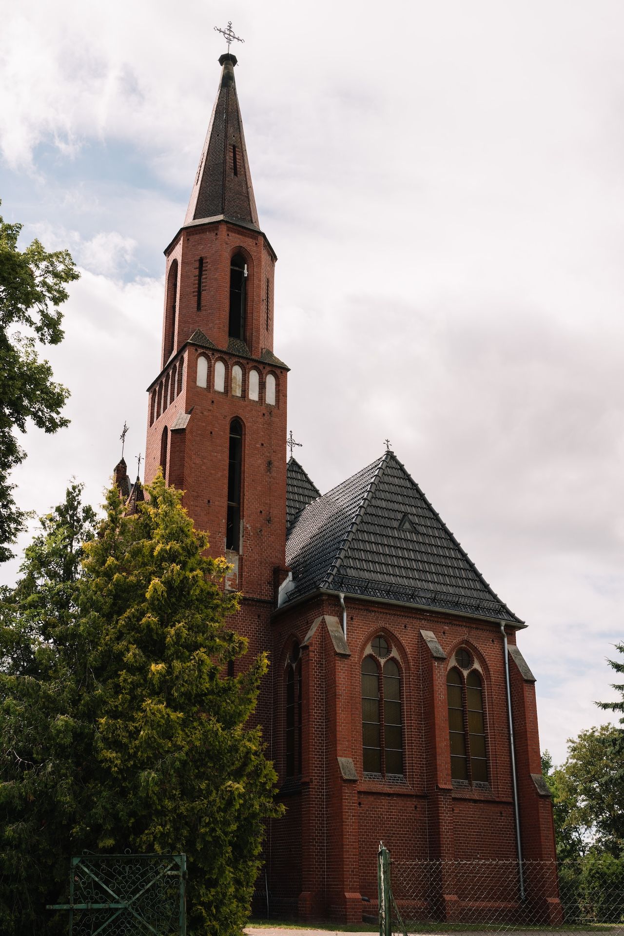 Red brick Gothic church with tall steeple and cross against cloudy sky exterior view.