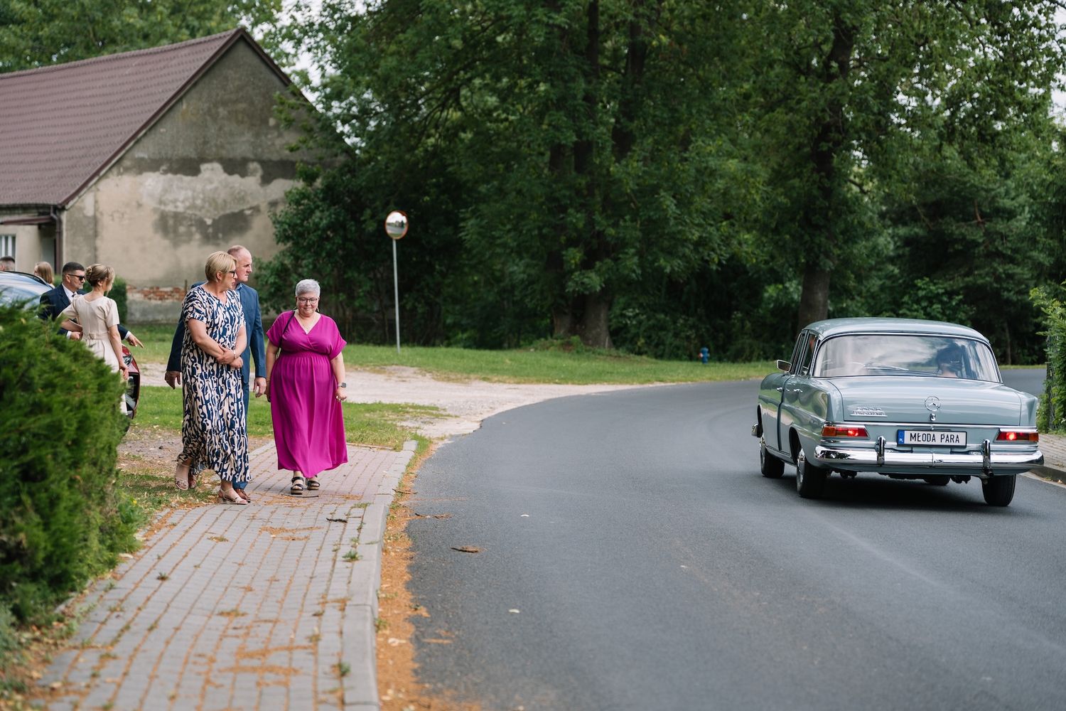 Elderly women in colorful dresses walk along brick path as vintage car passes on road.