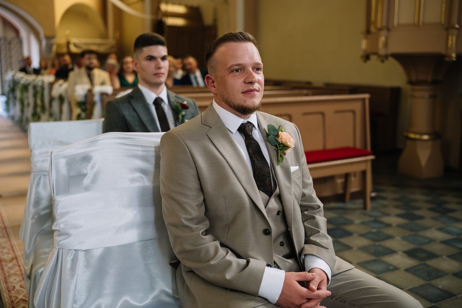 Groom in beige suit with boutonniere sits at church pew awaiting wedding ceremony.