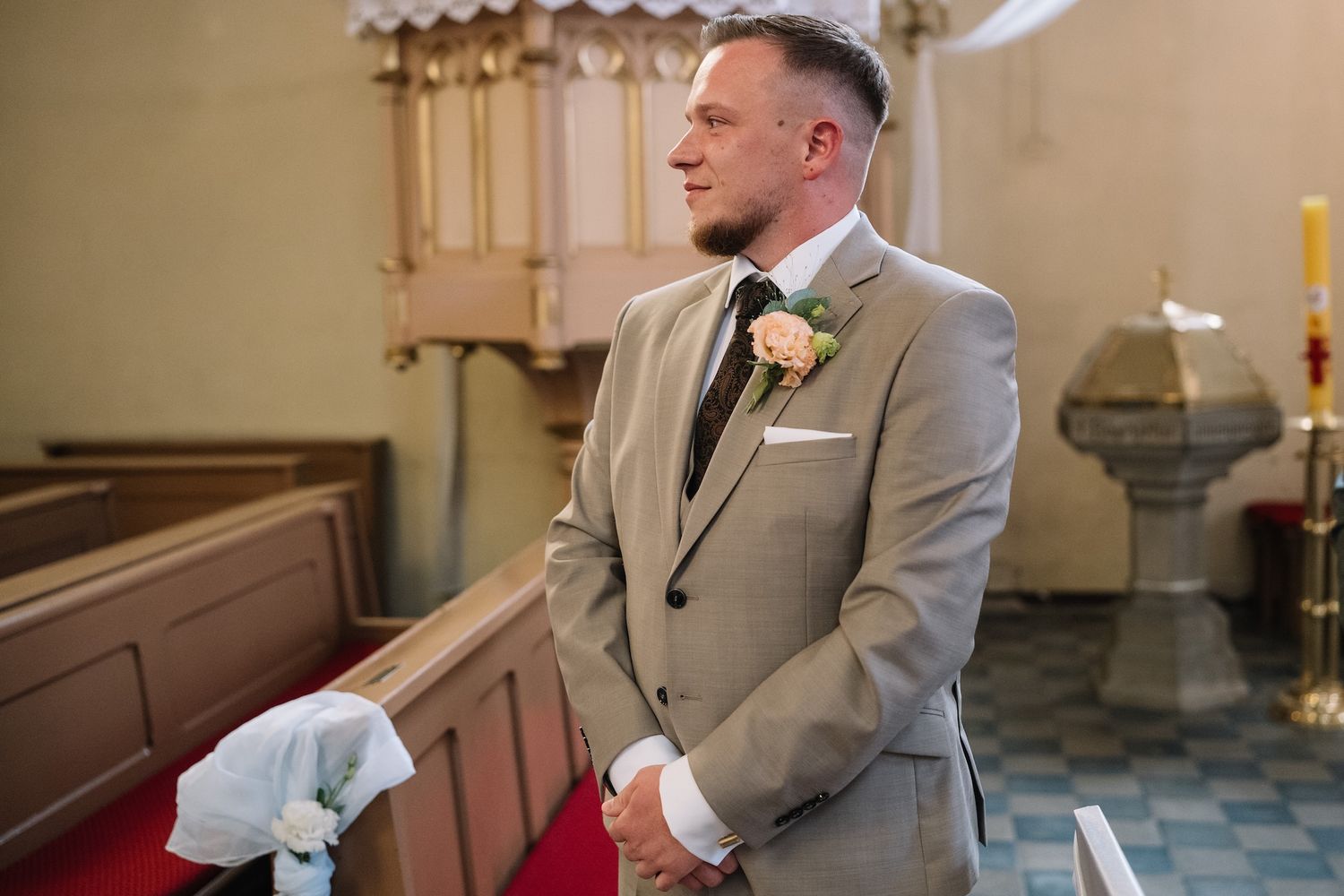 Groom in tan suit with floral boutonniere stands solemnly at church altar awaiting bride.