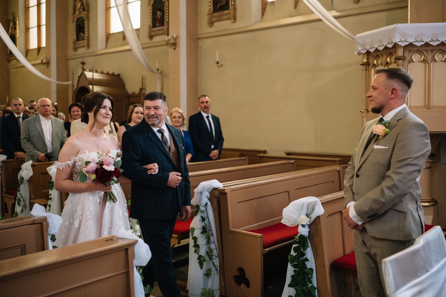 Bride in white gown walks down church aisle escorted by father toward waiting groom.