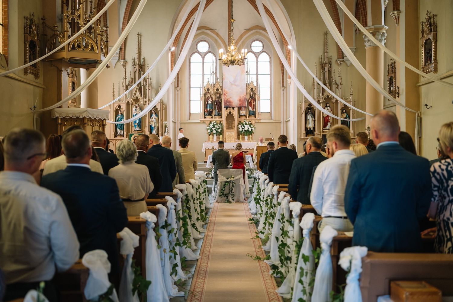 Guests fill wooden pews during elegant church wedding ceremony decorated with white draping.