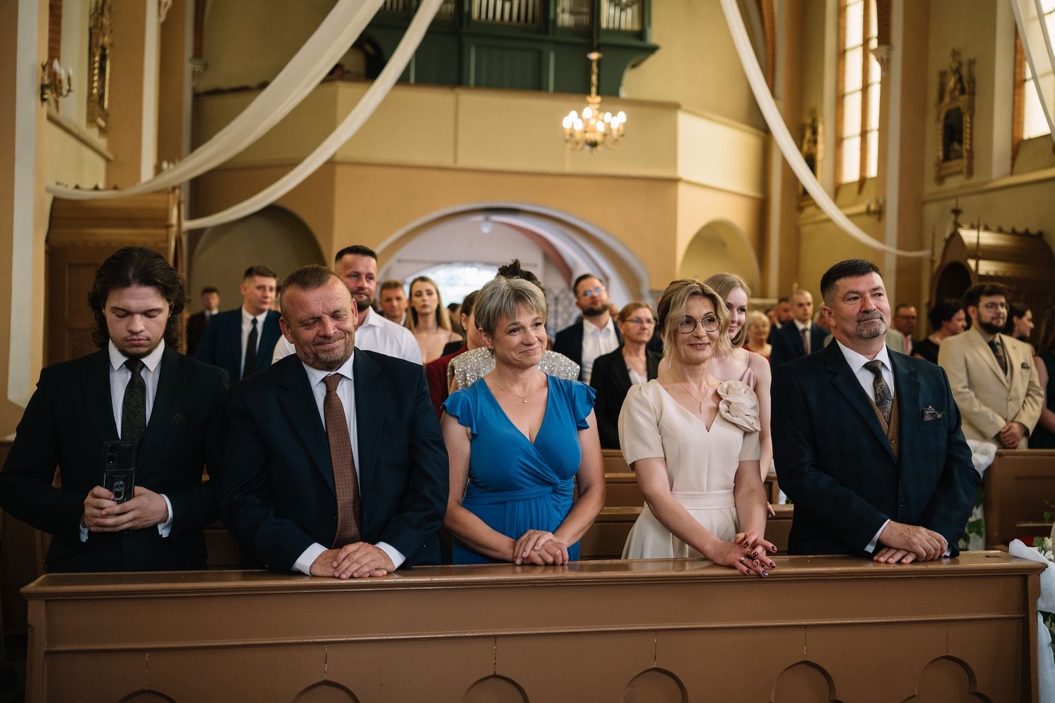 Wedding guests sit attentively in church pews during ceremony, dressed in formal attire.