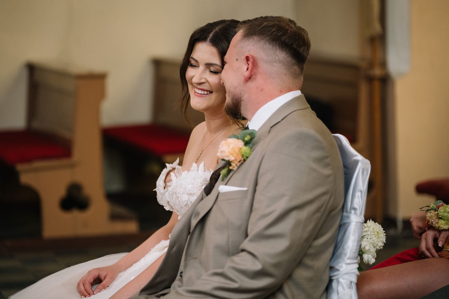 Bride in white dress smiles joyfully at groom in tan suit during church ceremony.
