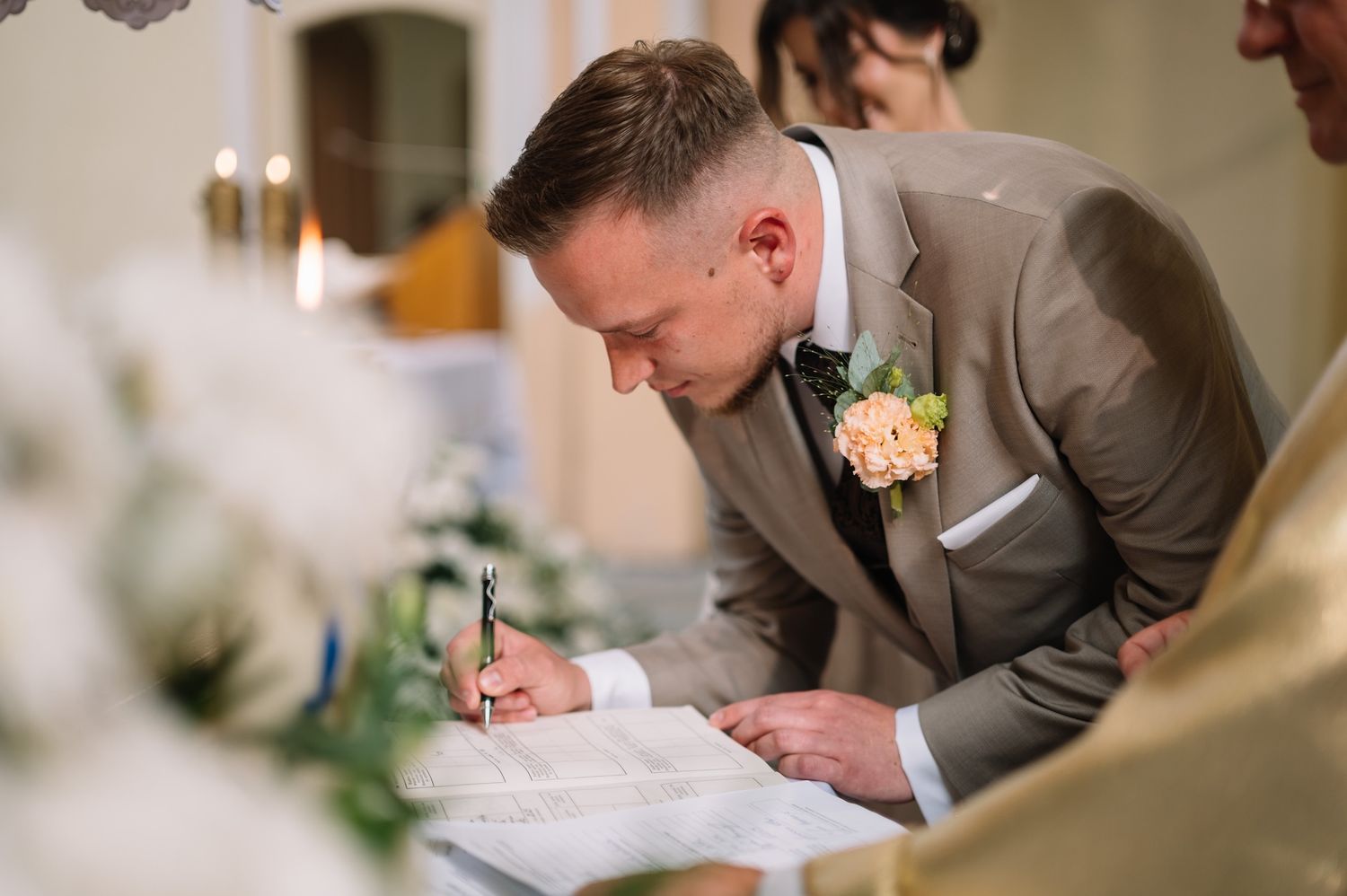 Groom in beige suit signs official wedding documents at church altar during ceremony.