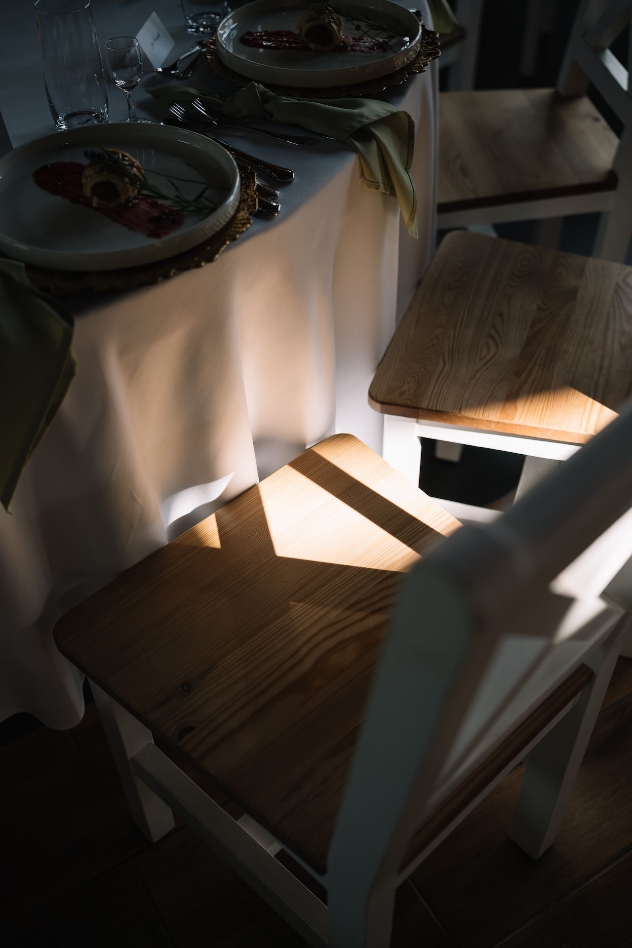 Elegant wedding reception table with white linen, fine dining plates, and wooden chairs illuminated by warm natural light.