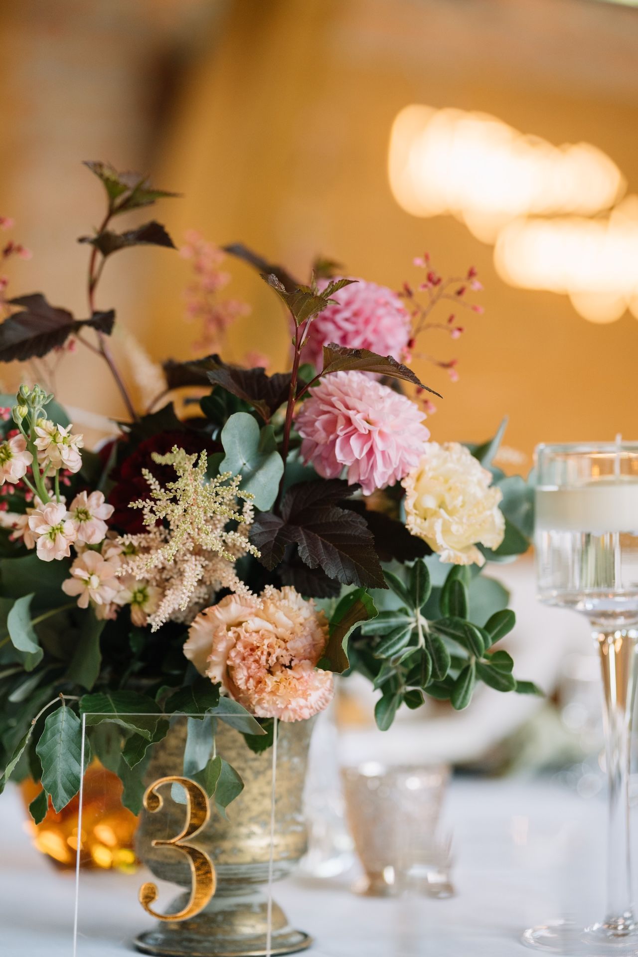 Beautiful wedding centerpiece with pink dahlias, peach carnations, and lush greenery beside a gold table number 3.