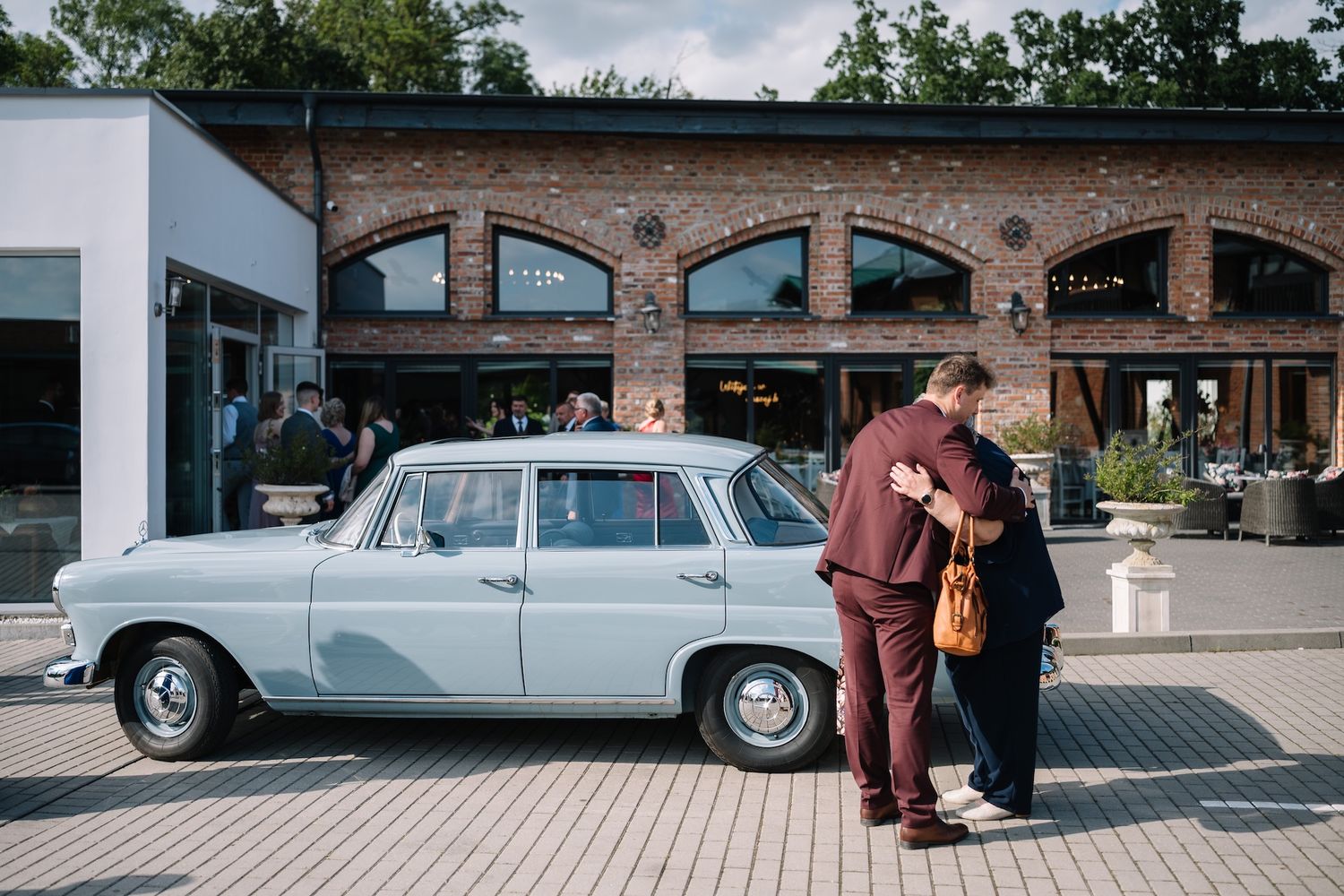 Couple embracing beside a classic vintage light blue car outside a brick wedding venue.