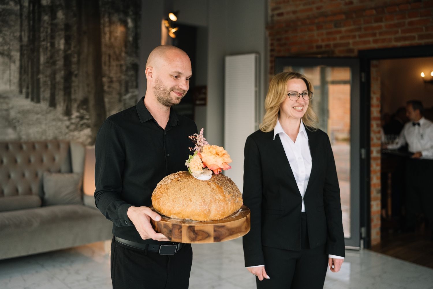 Smiling staff presenting a decorative rustic bread loaf on a wooden board at a wedding venue reception.