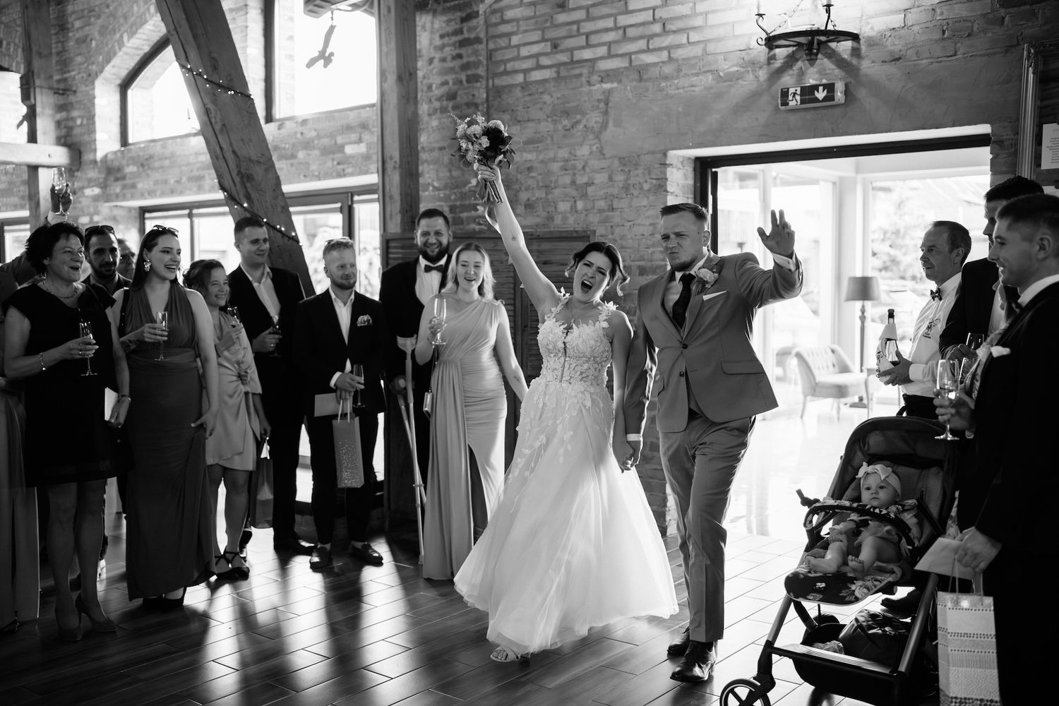 Black and white photo of bride and groom making grand entrance to cheering wedding guests in rustic barn venue.