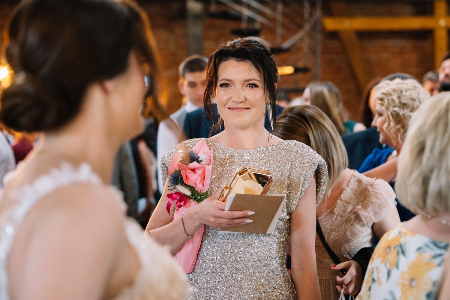 Smiling wedding guest in silver sequin dress holding pink flowers and a gold gift box at a wedding reception.