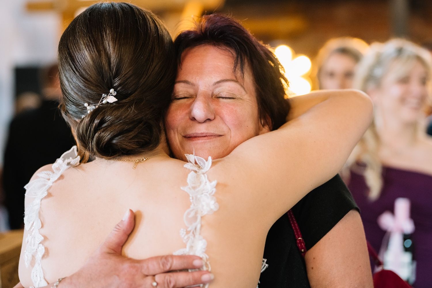 Emotional mother hugging bride in lace wedding dress at a rustic barn wedding reception.