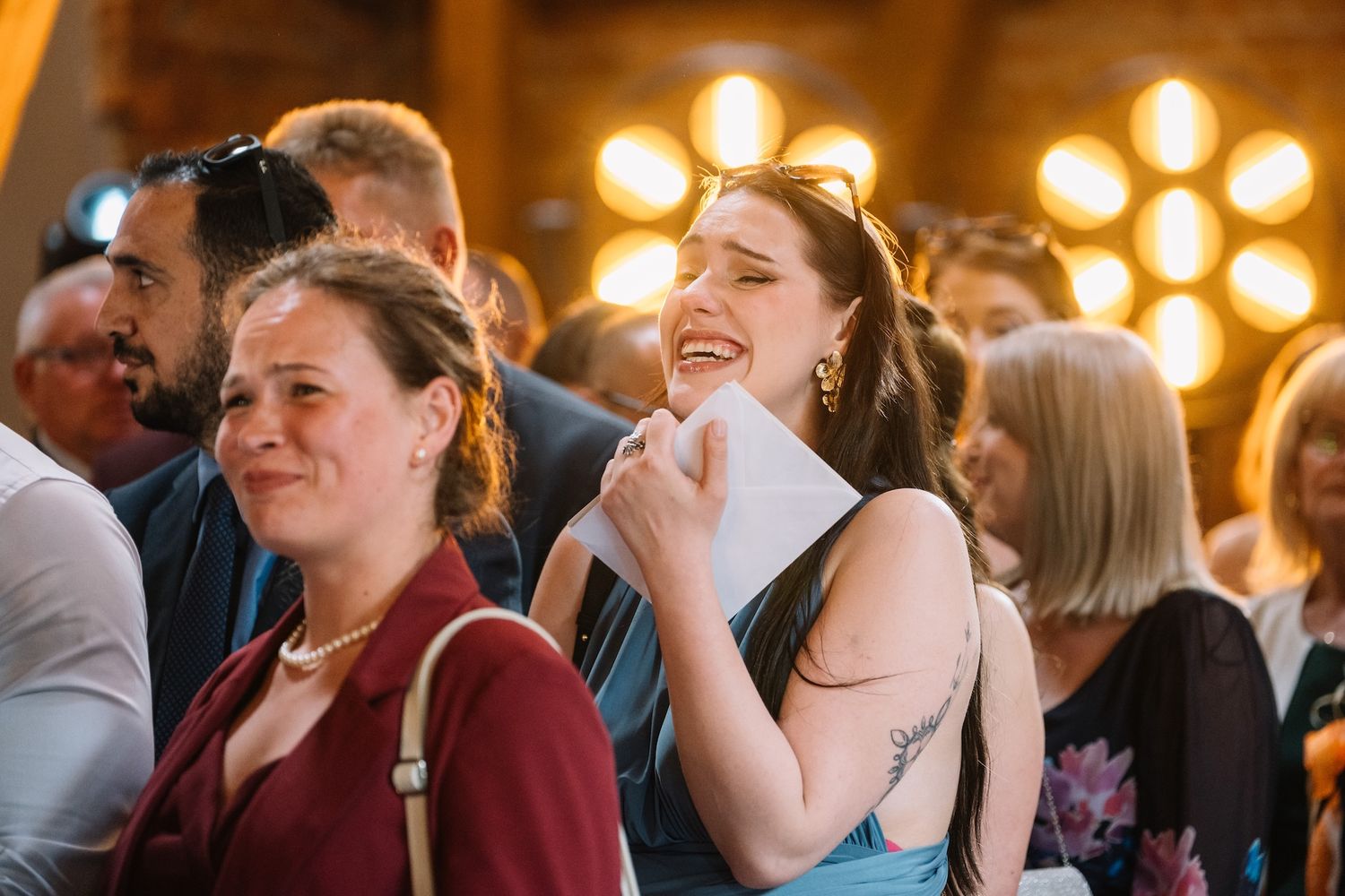 Joyful wedding guest laughing and holding a program at a lively indoor wedding reception.