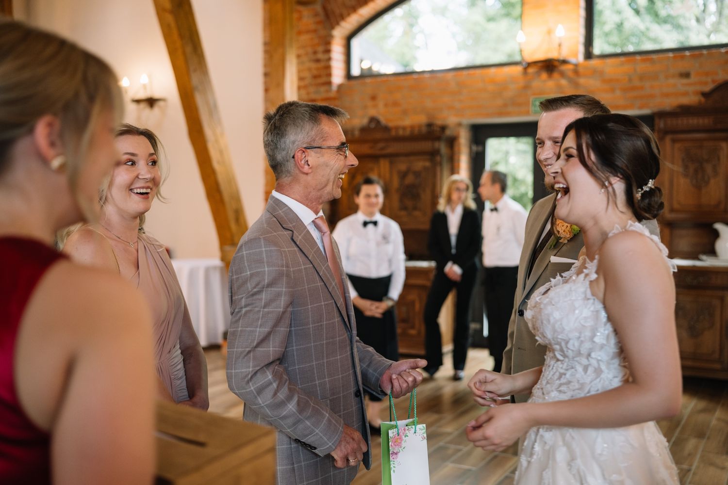 Bride in lace gown laughing with wedding guests in a warmly lit rustic brick reception venue.