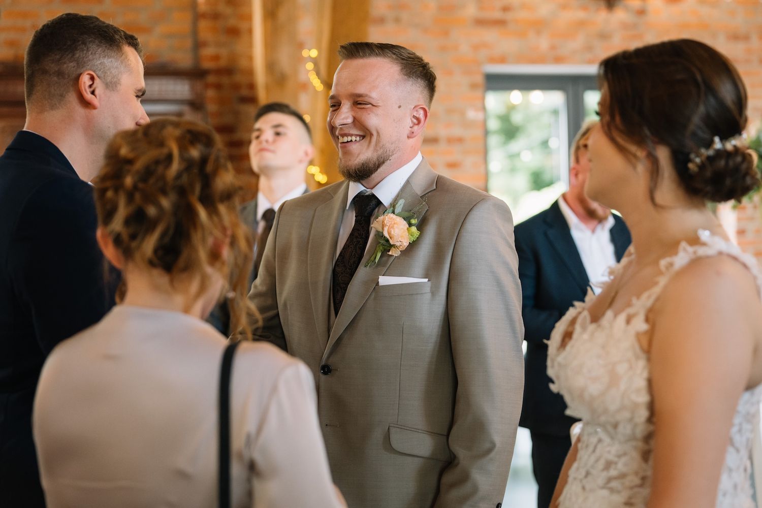 Smiling groom in beige suit with boutonniere greeting wedding guests at elegant brick venue ceremony.