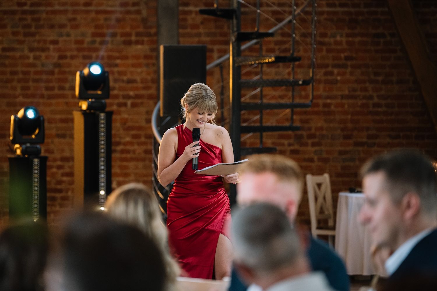 Elegant woman in red dress delivering speech with microphone at wedding reception in brick venue.
