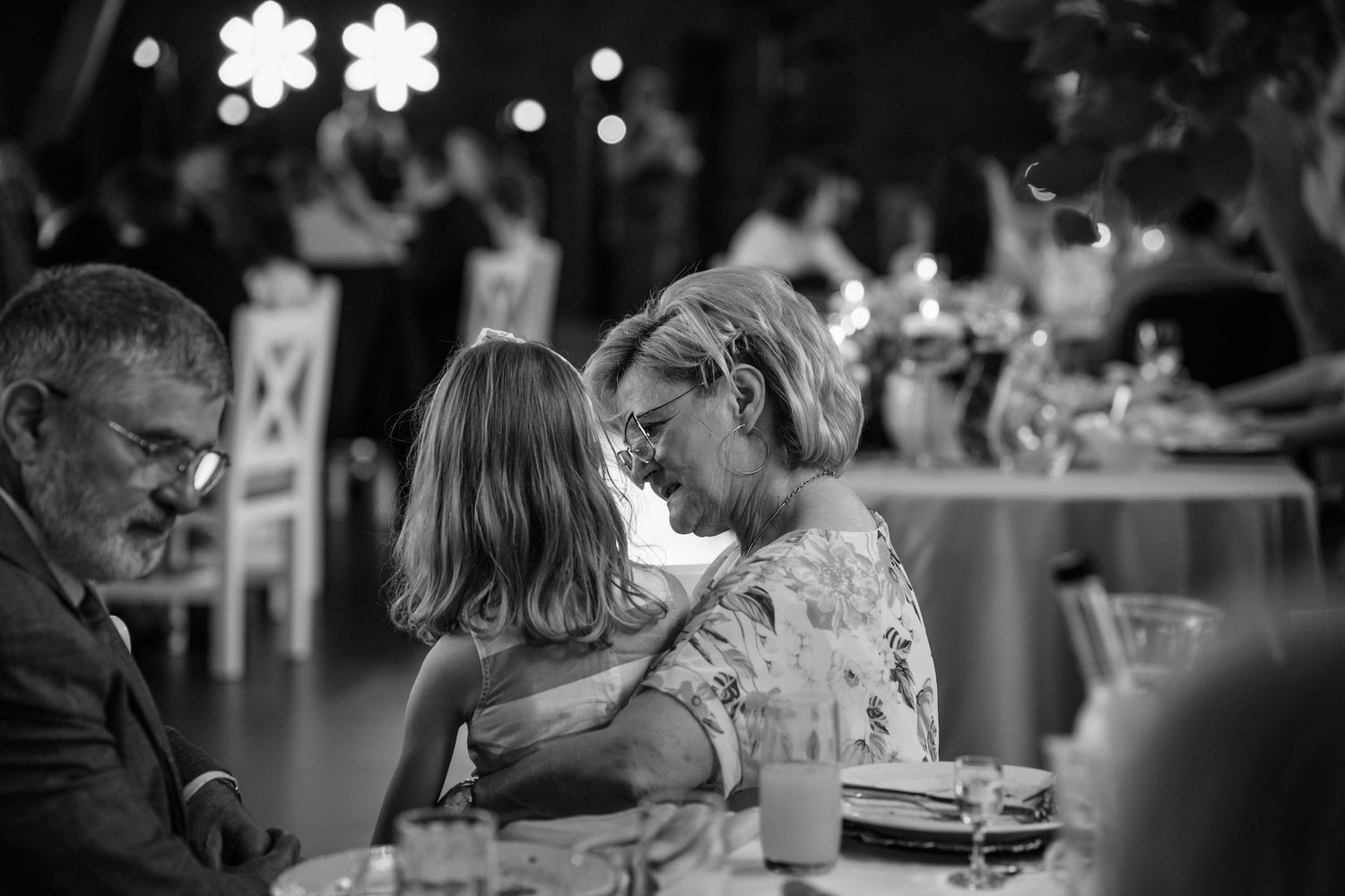 Tender black and white moment of grandmother embracing young child at wedding reception dinner table.