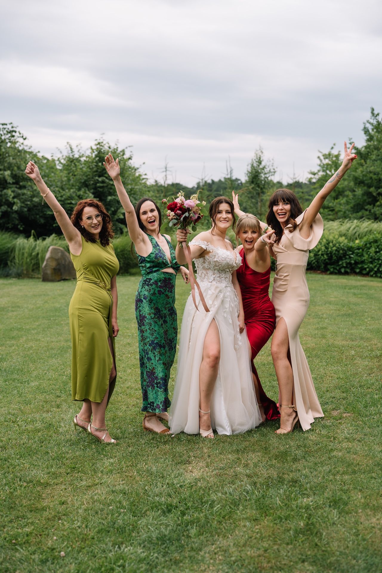 Joyful bride in lace gown celebrating with four bridesmaids in colorful dresses on green lawn.