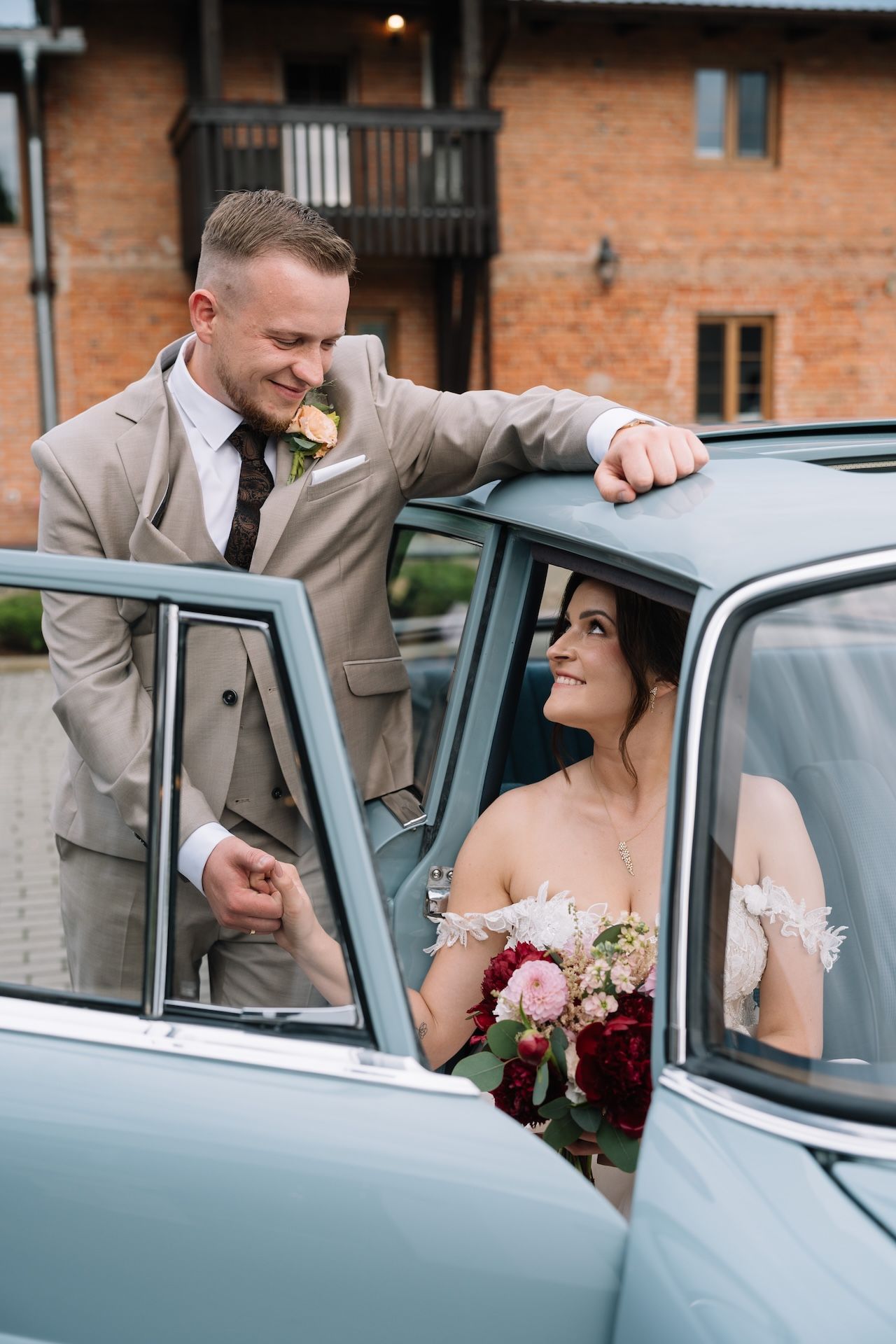 Groom in beige suit smiling at bride seated in vintage light blue classic wedding car.