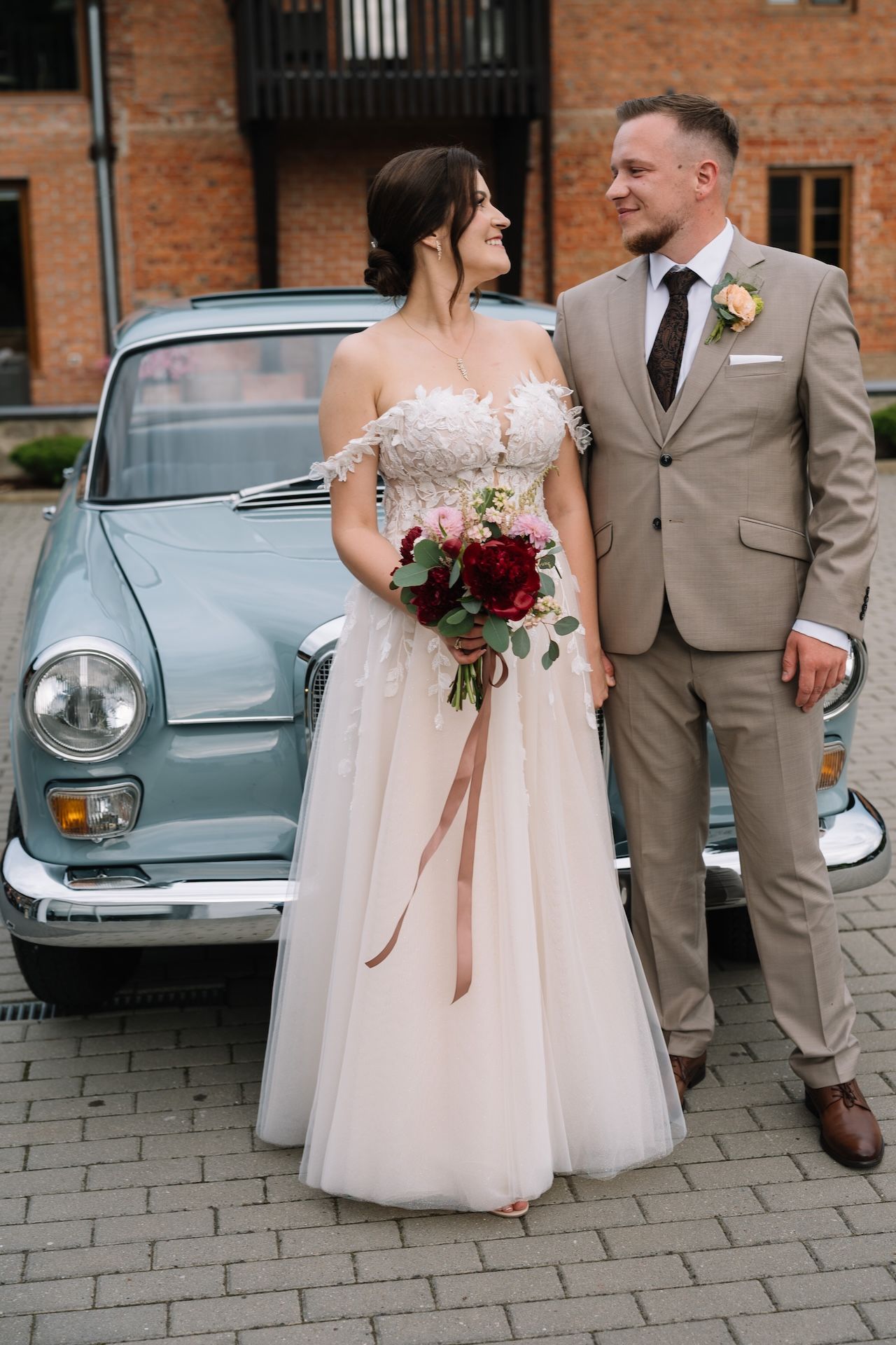Bride and groom sharing romantic moment beside vintage light blue Mercedes at brick building venue.