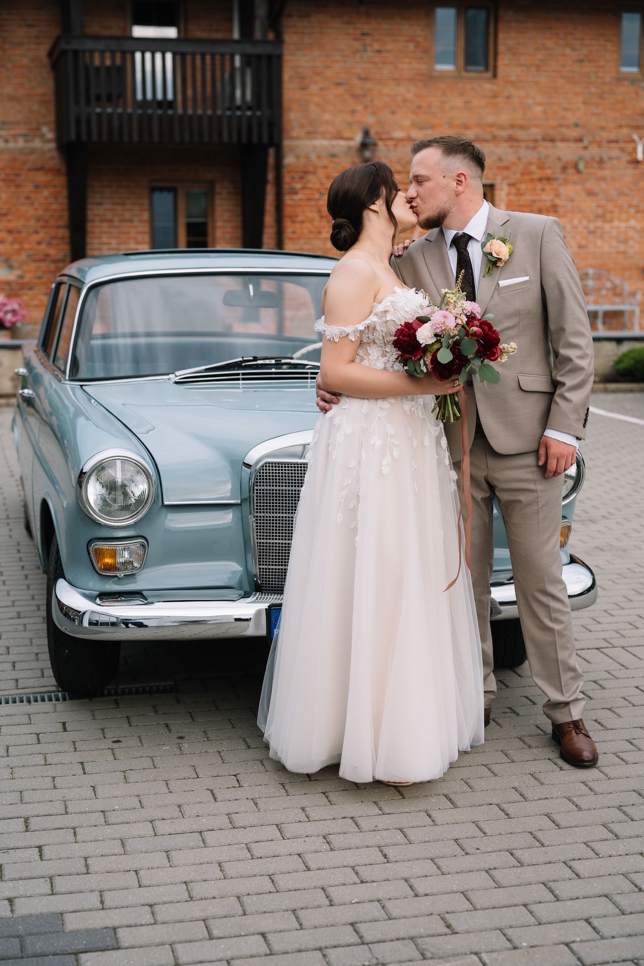Bride and groom sharing a kiss beside a vintage light blue Mercedes at rustic brick venue.