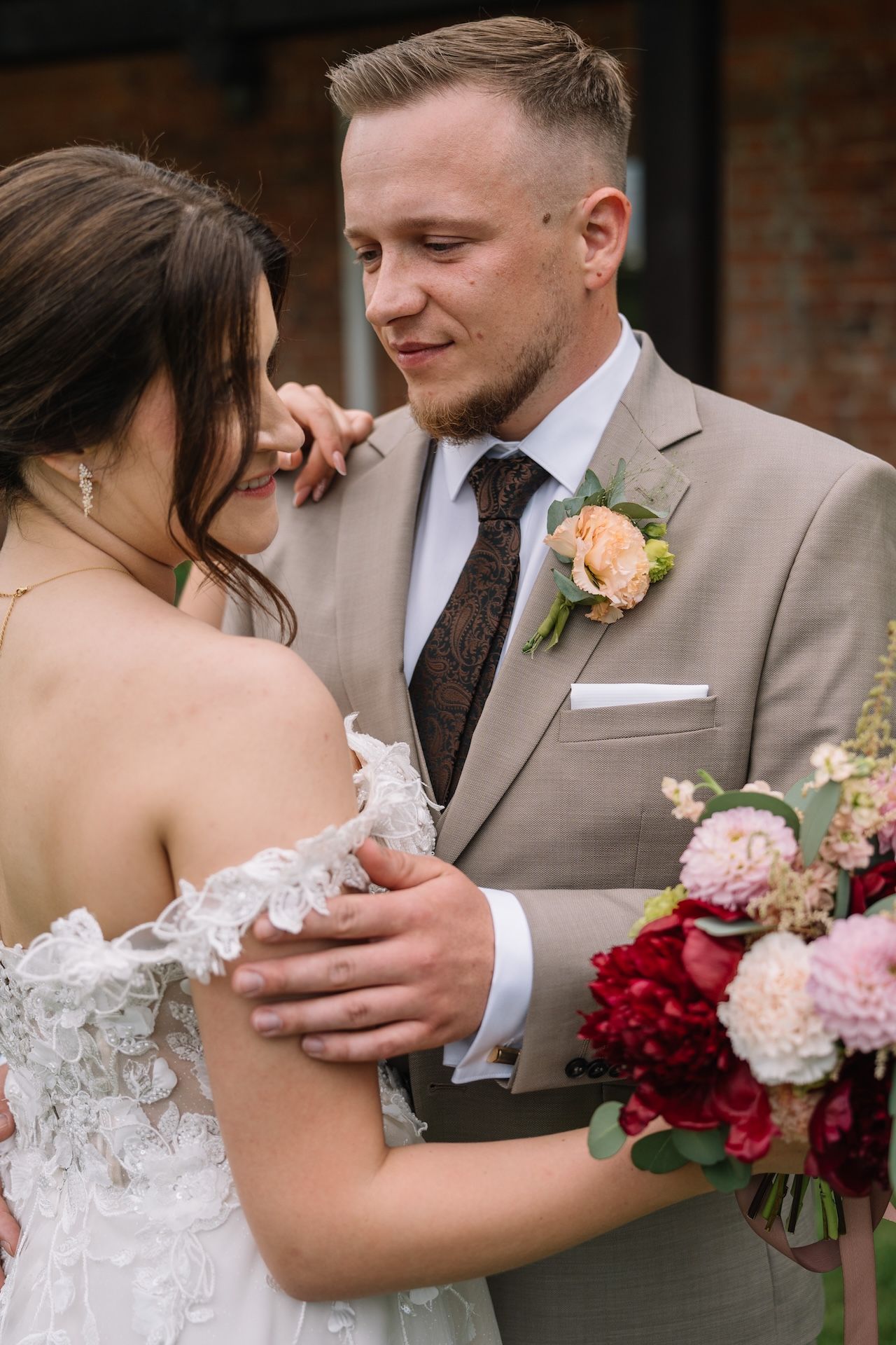 Groom in beige suit tenderly embracing bride in lace off-shoulder gown holding colorful bouquet.