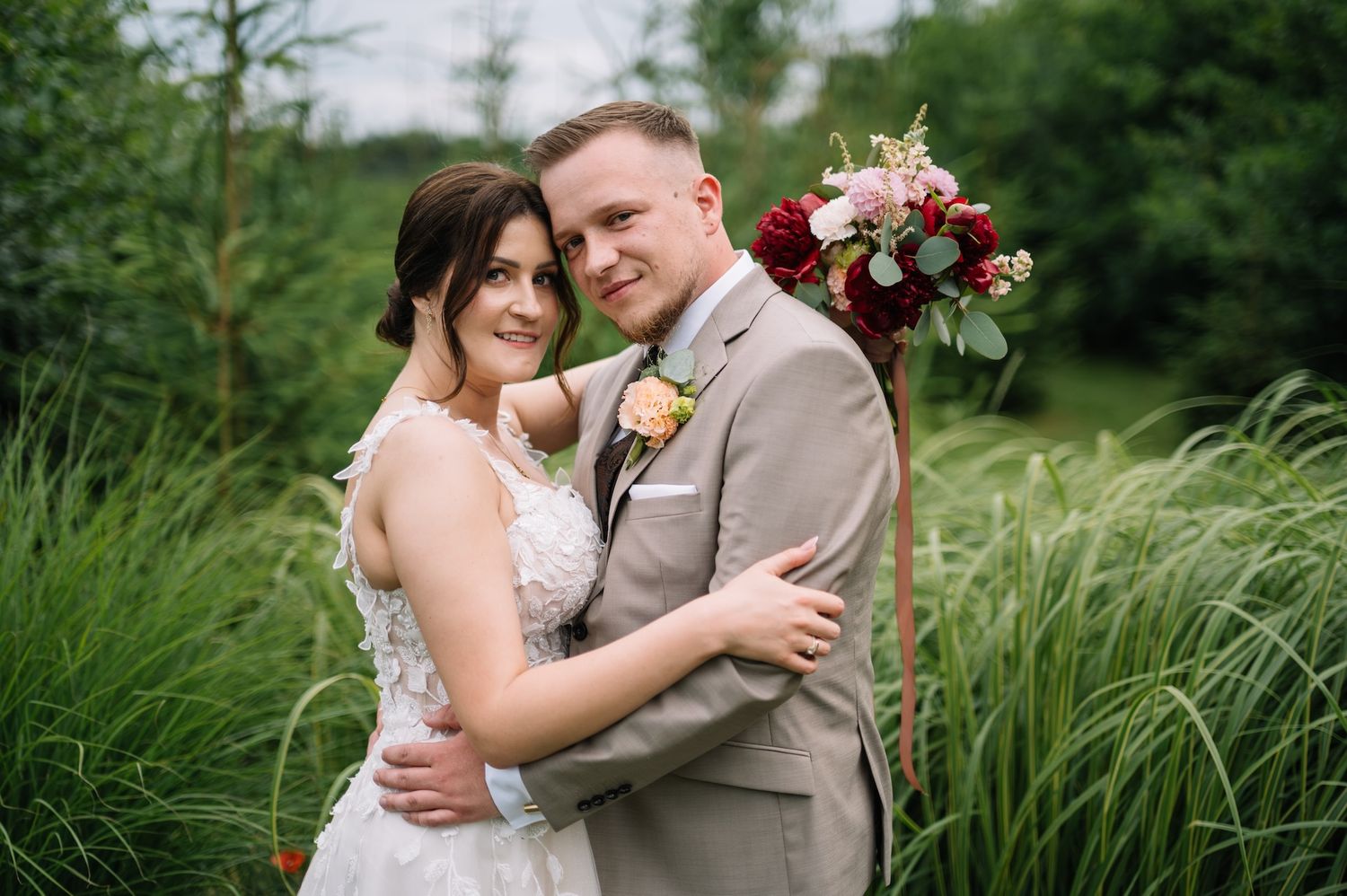 Bride and groom embracing in lush green garden, groom in tan suit with boutonniere.