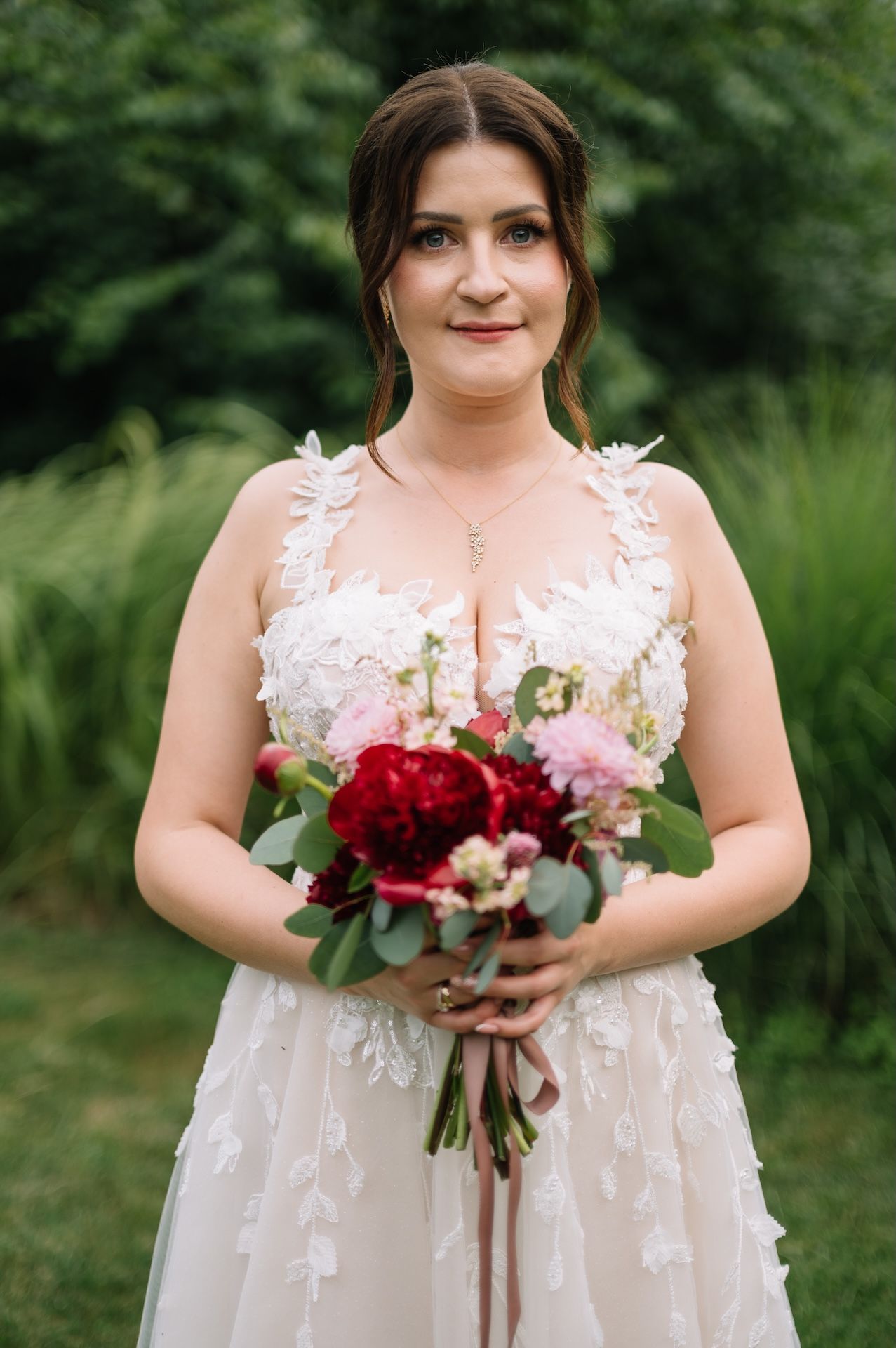 Bride in floral lace wedding gown holding red and pink bouquet outdoors.