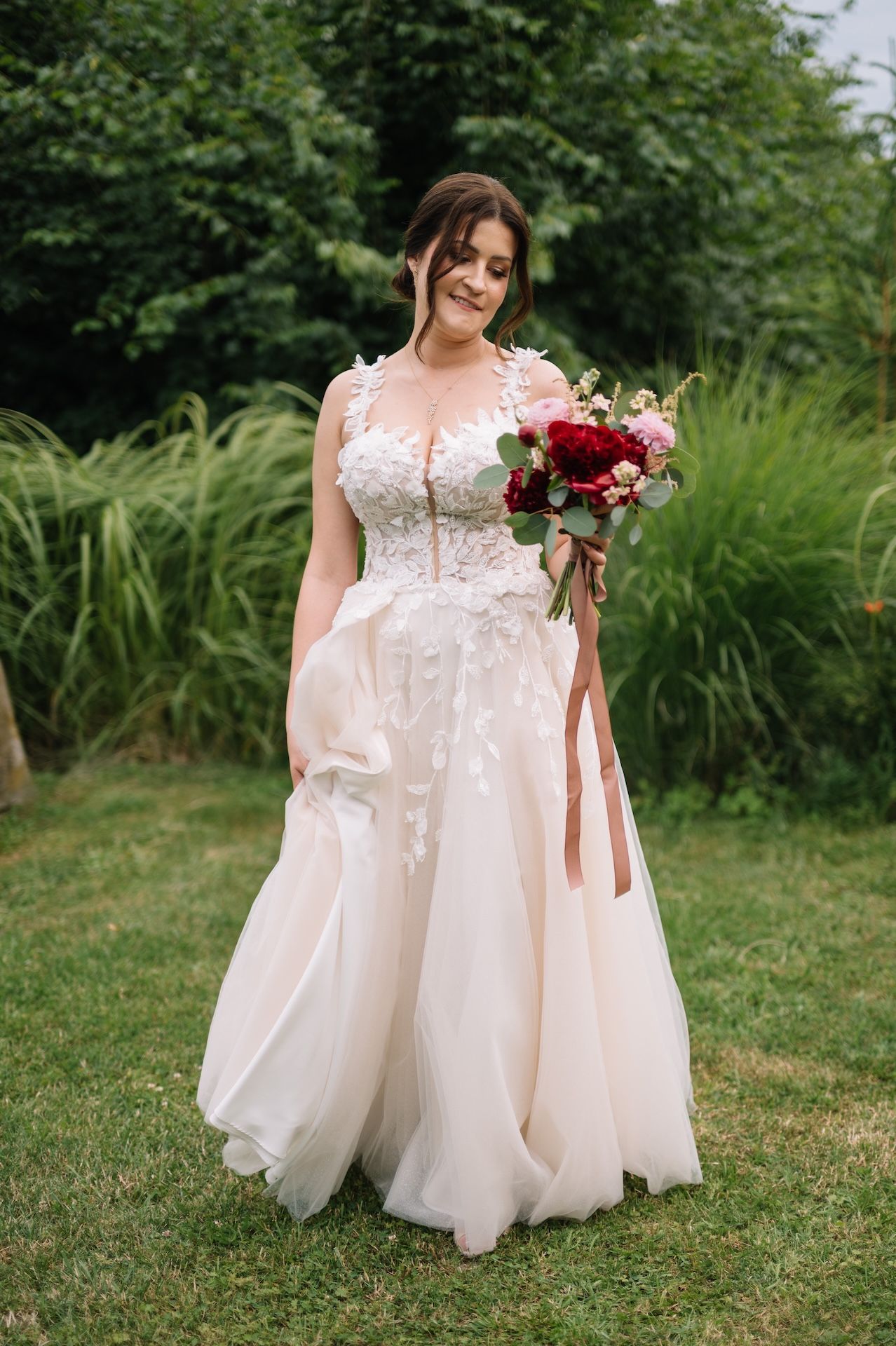 Bride in blush A-line wedding dress with floral appliques holding bouquet on green lawn.