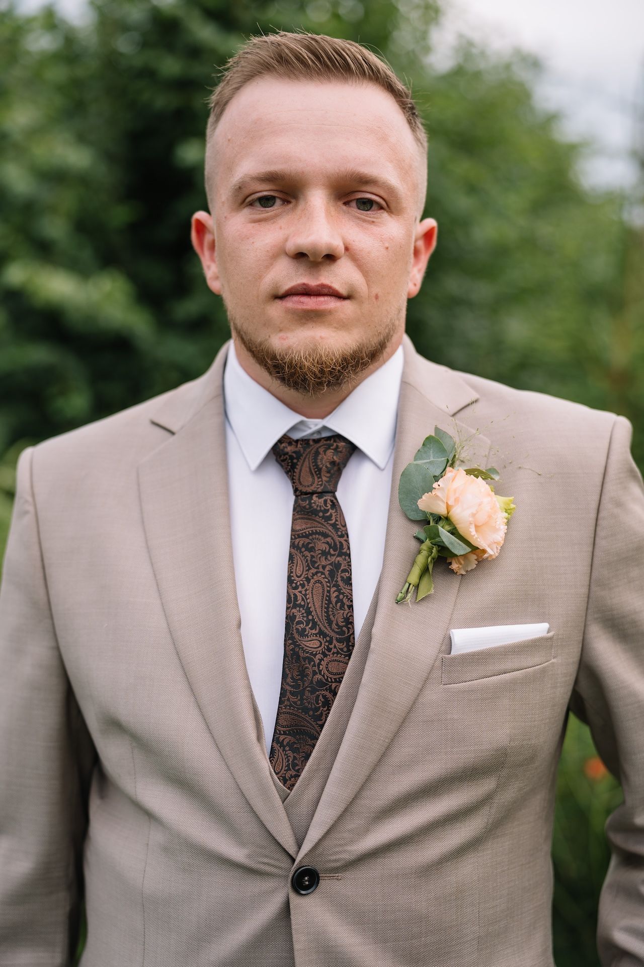 Groom in beige suit with paisley tie and peach boutonniere posing outdoors.