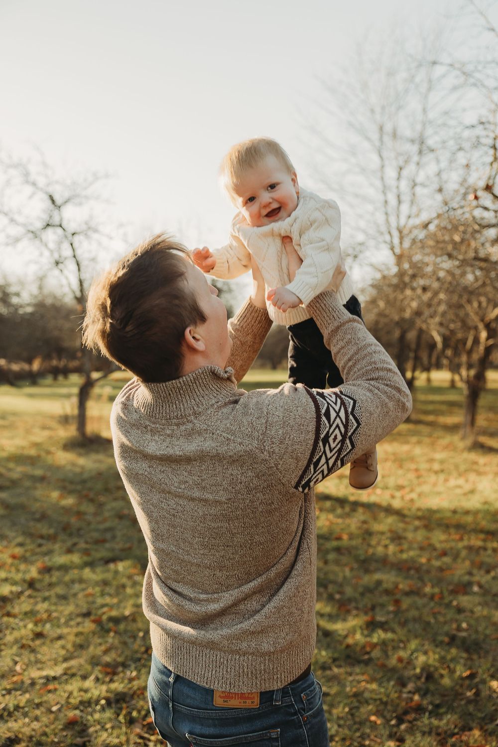 A parent in a knit sweater lifts a baby wearing white clothing outdoors during golden hour in an orchard setting.