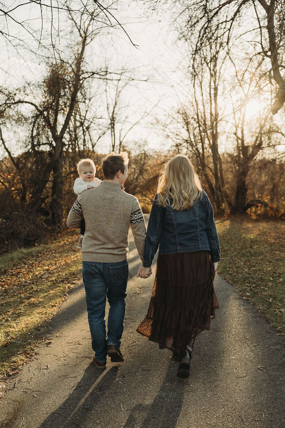 A couple walks hand in hand down a sunlit path through autumn trees casting long shadows at sunset.