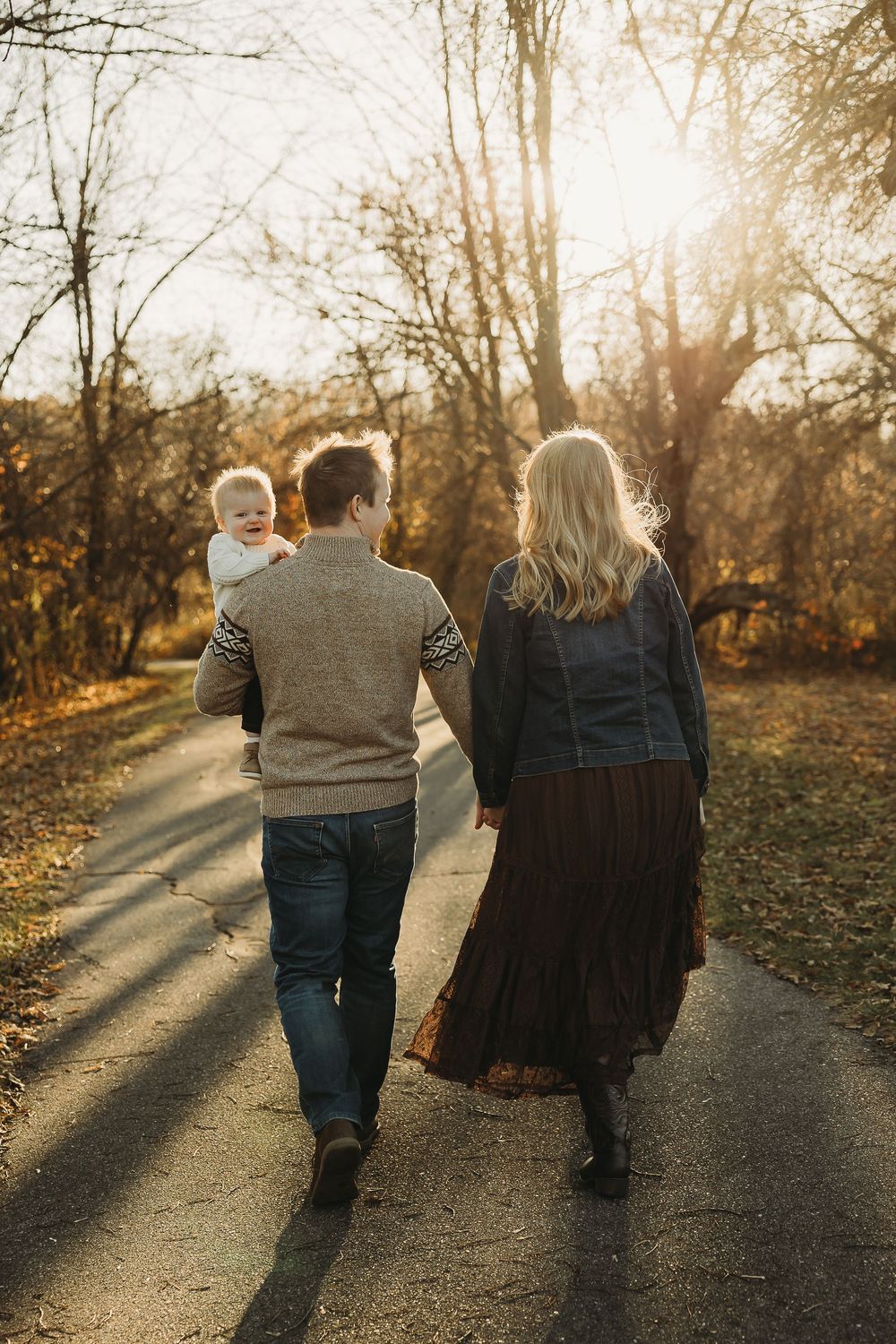 Two people walk hand in hand down a sunlit autumn path as golden light filters through bare trees.