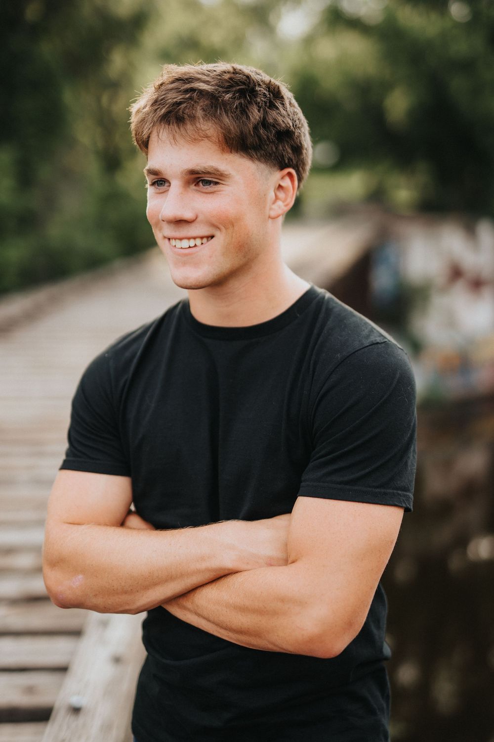Young man in black t-shirt stands with arms crossed and smiles while posing outdoors in natural lighting.