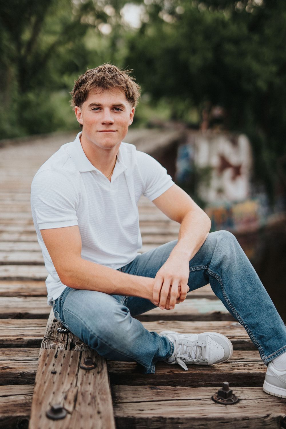 Person in white polo shirt and jeans sits casually on wooden surface with urban background.