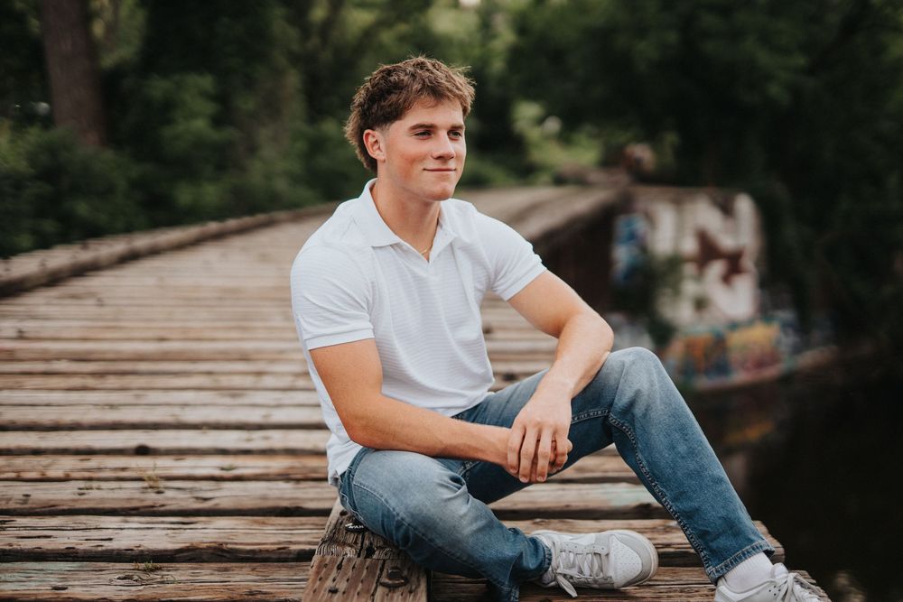 Young man in casual attire sits on wooden boardwalk during outdoor photo session at sunset.