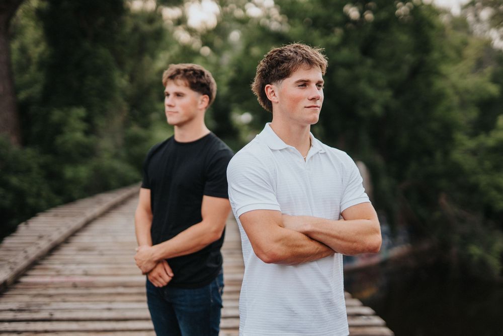Two people in black and white shirts stand on wooden planks in a lush outdoor setting.