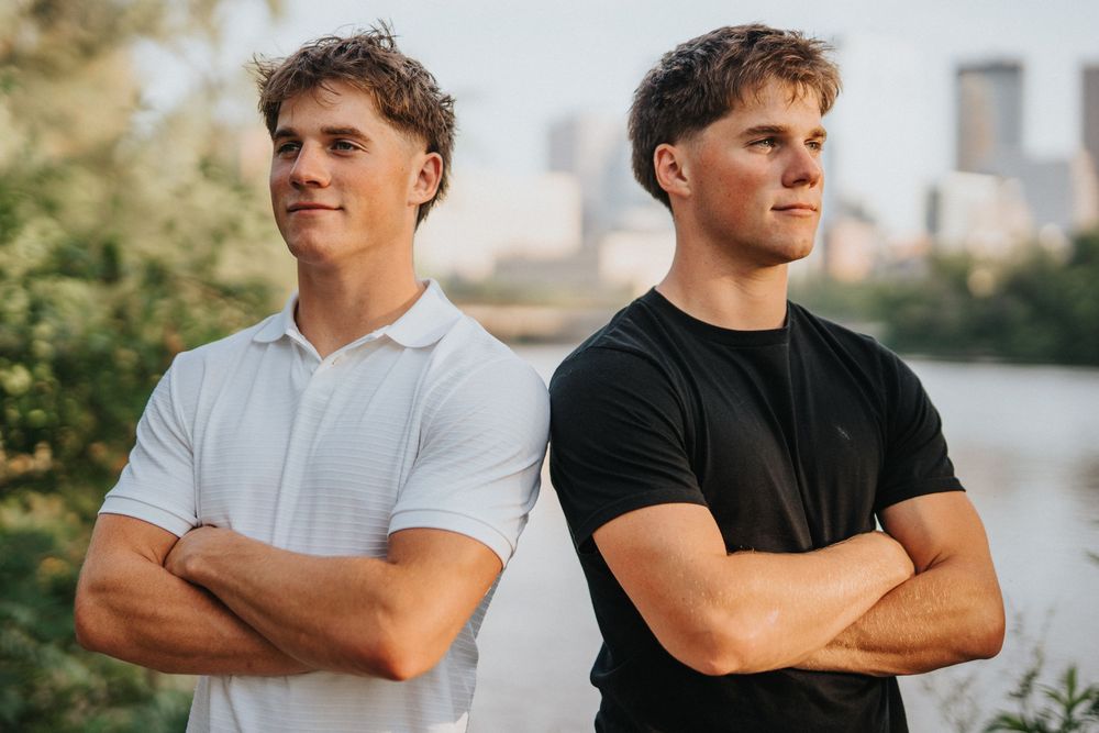 Two people stand side by side with arms crossed against a sunset cityscape backdrop.