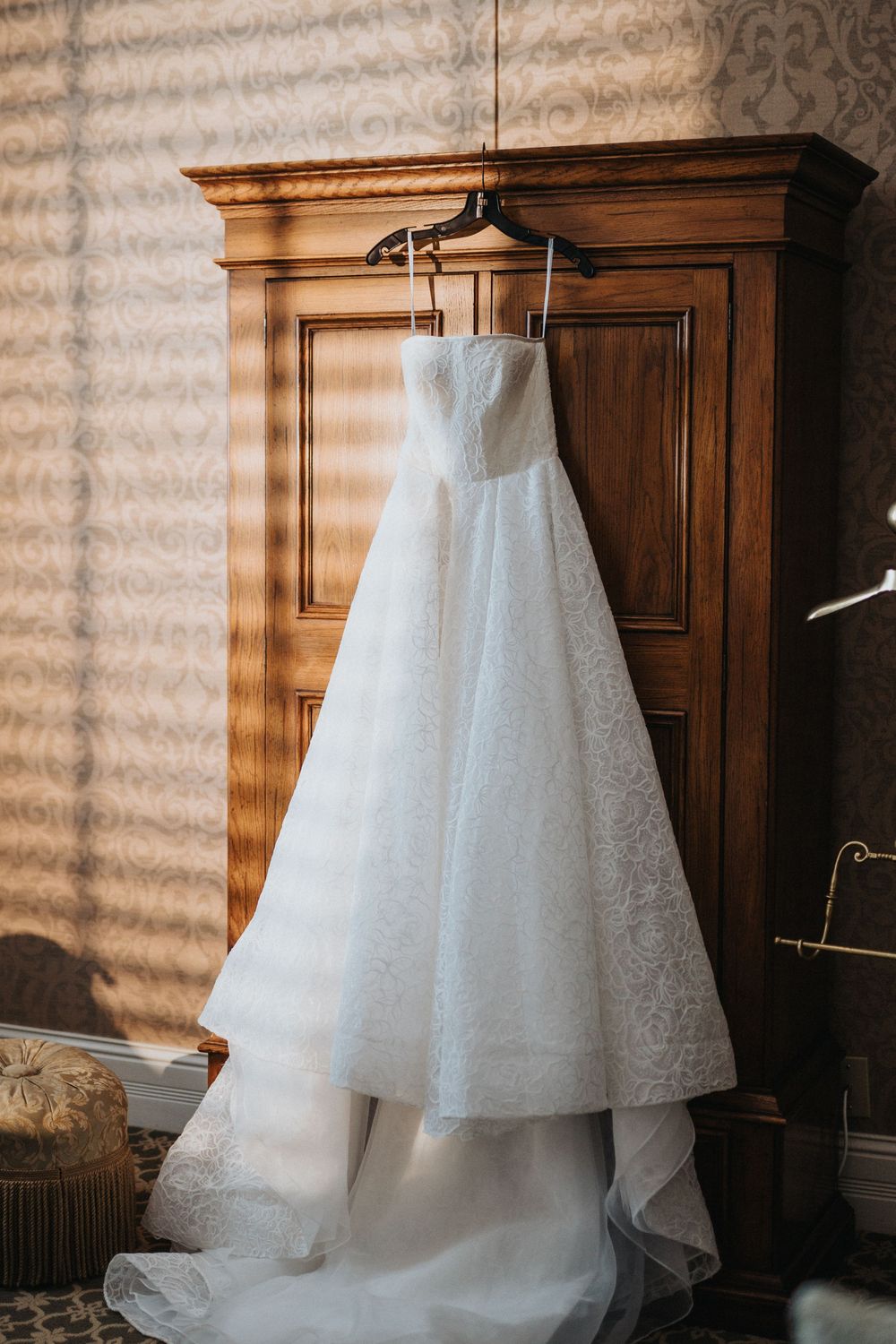 A flowing white wedding dress with delicate lace details hangs elegantly against a wooden wardrobe in warm lighting.