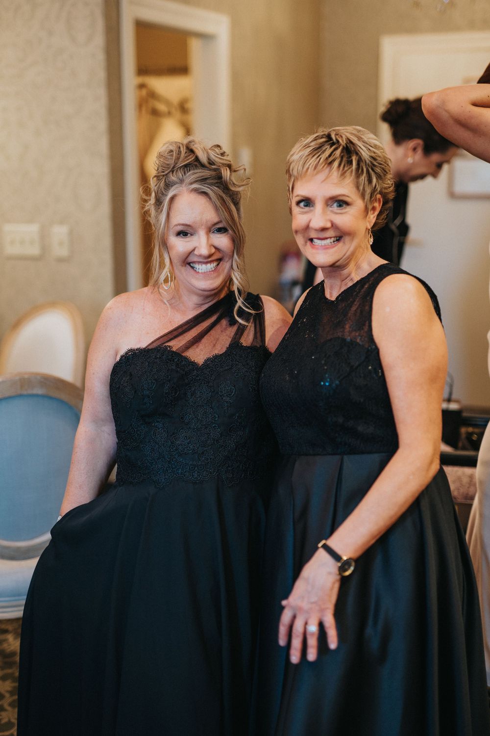 Two women wearing matching black dresses share a joyful moment during a wedding preparation.