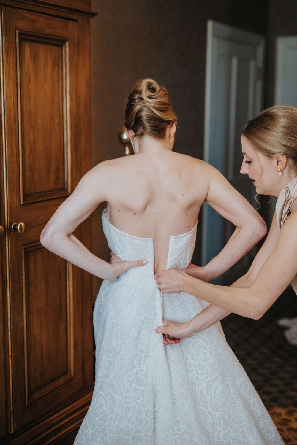 Multiple people help zip and adjust a white lace wedding dress during bridal preparations.