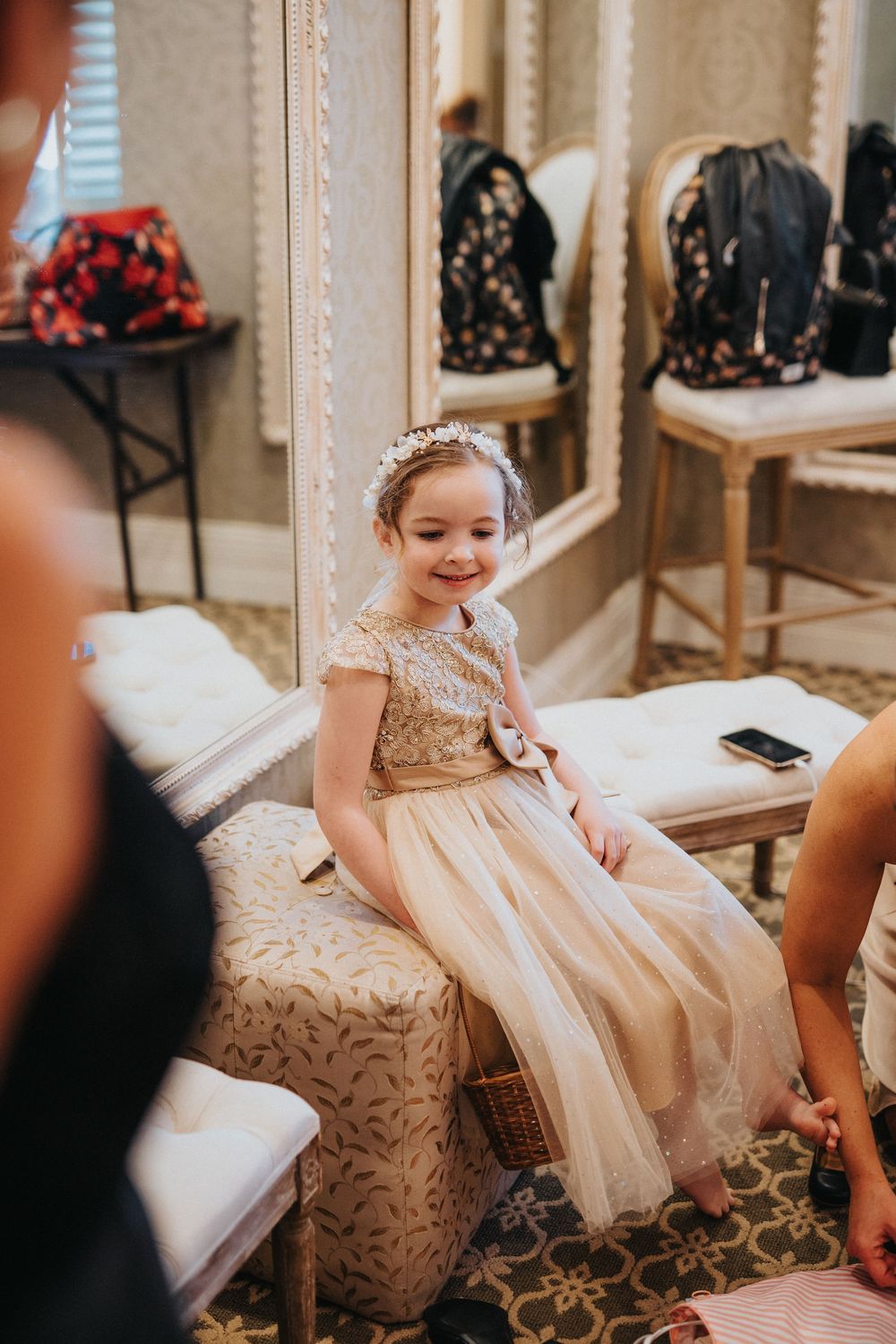 Young child in a white dress sitting on an elegant chair with decorative upholstery during a formal event.