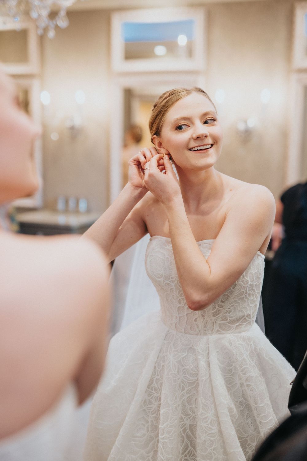 A person in a white lace wedding dress smiles and gestures while getting ready.